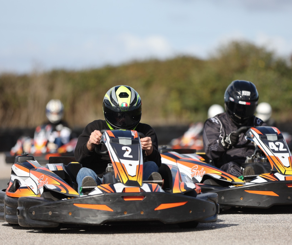 A group of people are mid-race at a go-kart track in the sunshine.