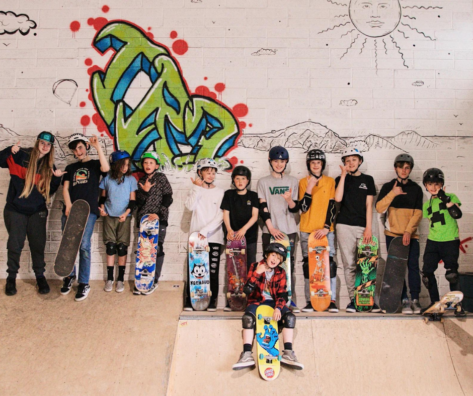 Photo of a group of children and teenagers at the top of a skateramp holiding multicoloured boards wearing helmets and brightly coloured clothing.