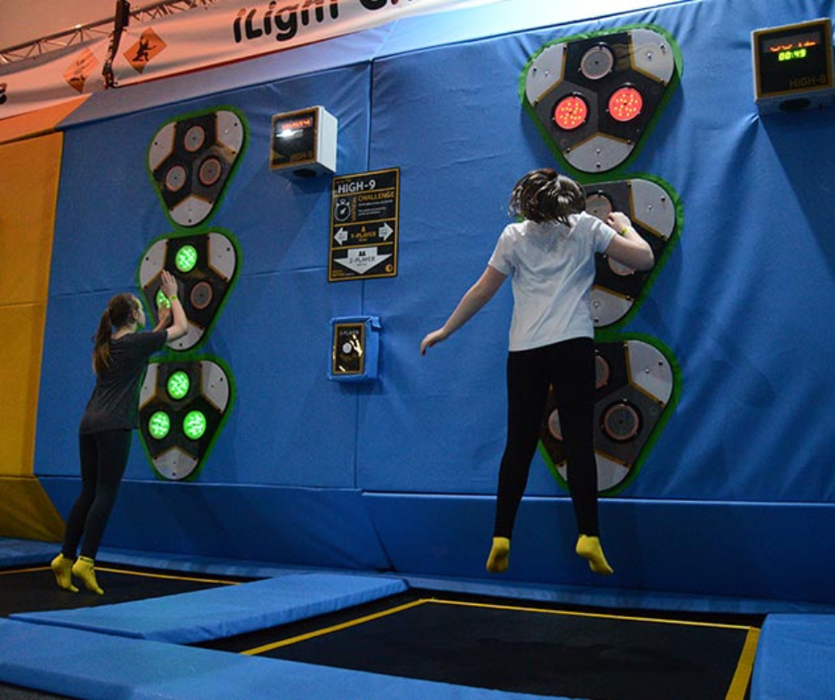Two children bouncing on a square trampoline. The child closest to the camera is mid-jump pressing a light-up reflex-test game.