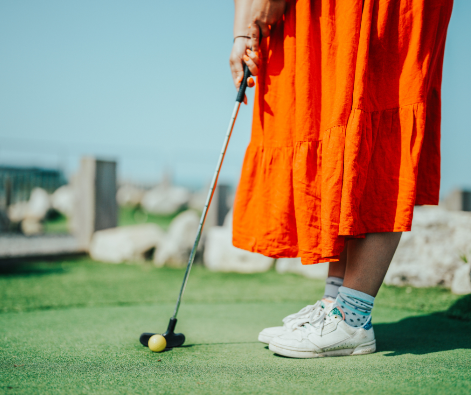 Photo of someone in a bright orange dress and white trainers putting a golf ball on a mini golf green with blue skies in the background.