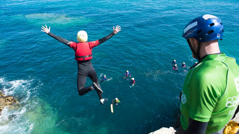 Photo of someone in a wetsuit and red lifejacket jumping off a cliff into the aqua blue sea on the coast of Cornwall