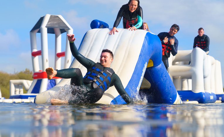 A group of young adults are laughing and climbing on an inflatable aqua park assault course. The man at the front of the photo is sliding down into the water whilst the others look on in amusement.