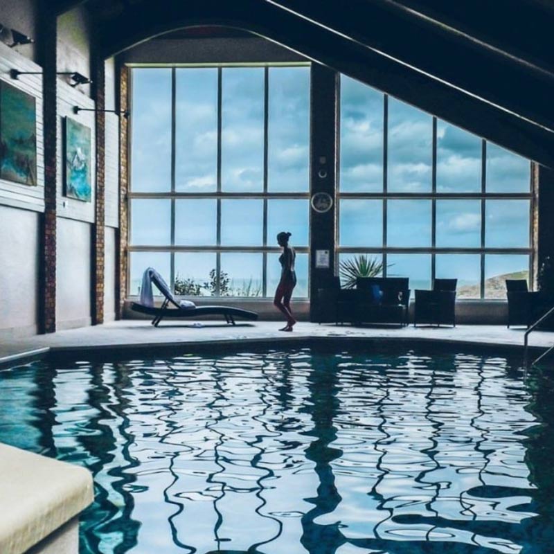 Indoor swimming pool with large windows overlooking the coast, featuring a person walking past lounge chairs. Spa pool at spa near Crantock Bay